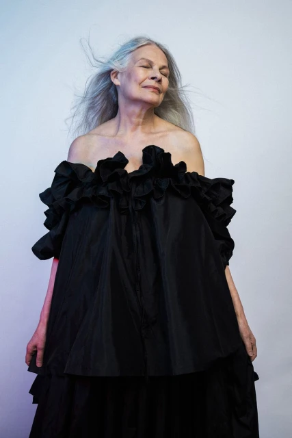 Elegant mature woman with grey hair poses in a black ruffled dress in a studio with blue and purple lighting. Fashion portrait.