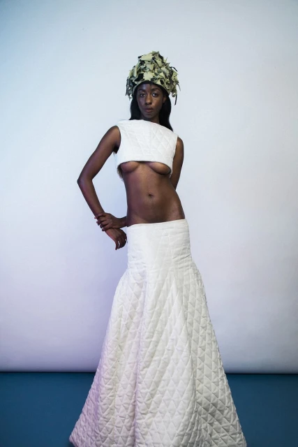 Woman in a white quilted two-piece outfit and leaf headdress poses in front of a gradient background in a studio shot.