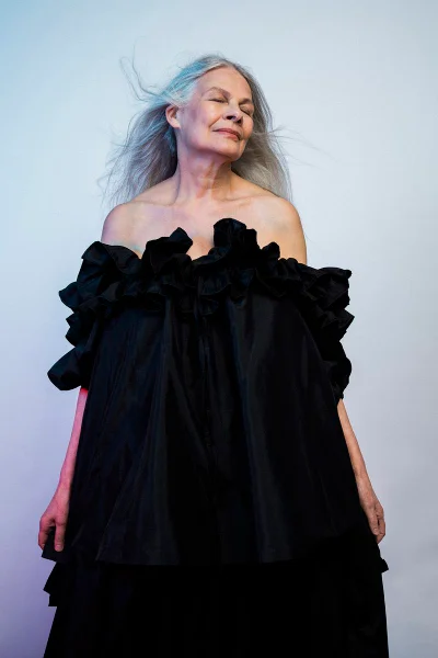 Mature woman with long silver hair posing in a voluminous black off-the-shoulder tiered ruffle dress against a studio backdrop.