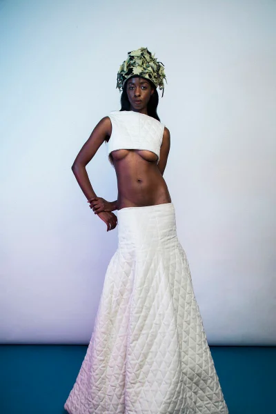 Model wearing a white quilted crop top, matching floor-length skirt, and a foliage headpiece against a pale blue studio backdrop.
