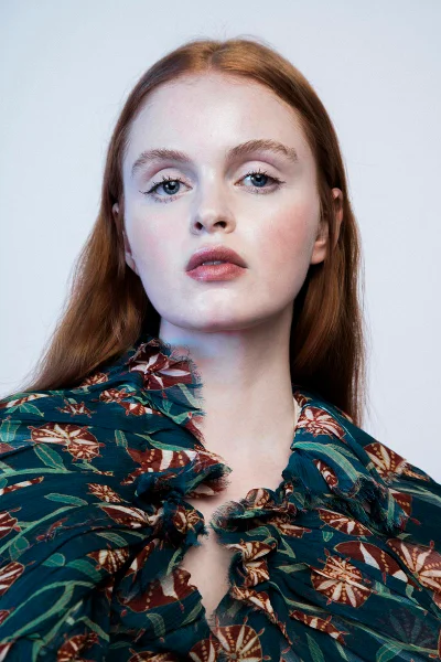 Portrait of a red-haired woman wearing a ruffled botanical print teal garment against a neutral pale studio backdrop.