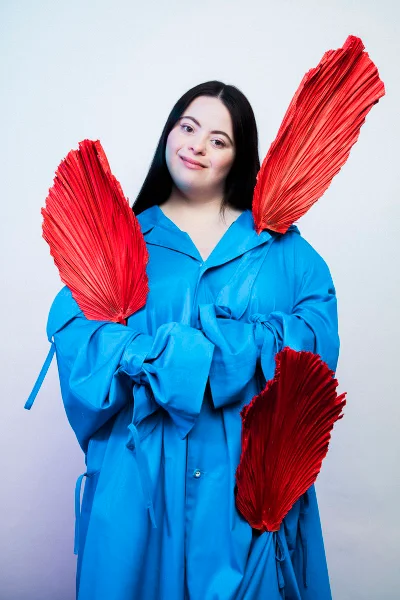 Model Ellie Goldstein smiling in an oversized bright blue coat surrounded by red pleated palm leaf fans against a studio background.