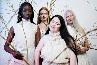 Four female models wearing white garments posed together behind slender bare branches against a white studio background.