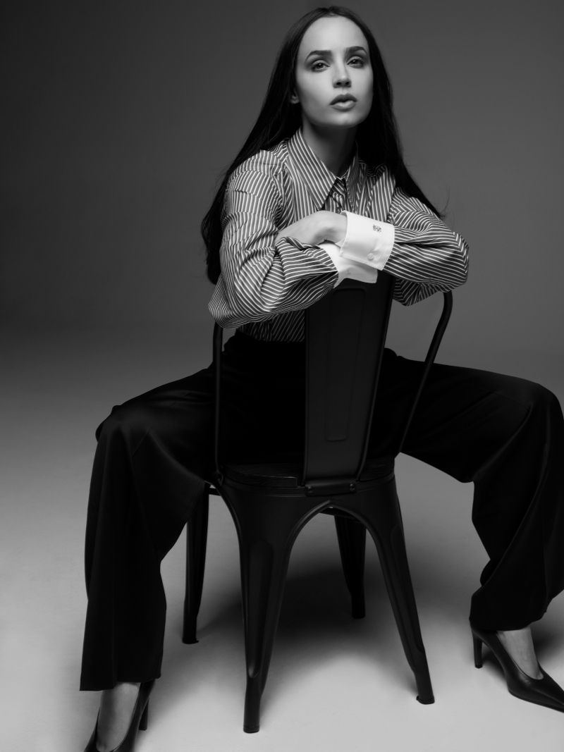 Elegant woman in a striped shirt and wide-leg pants sitting backwards on a metal chair in a monochrome studio shot.