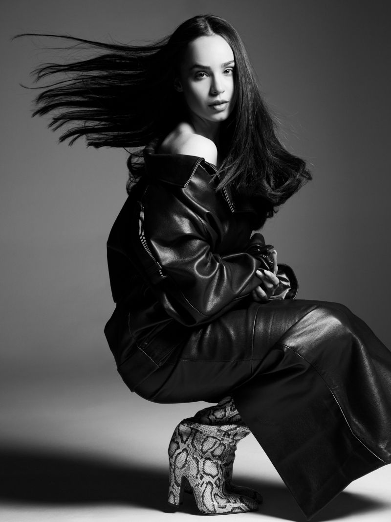Monochrome portrait of a woman in leather jacket and pants, snakeskin boots, with hair blowing, posing in a photo studio.