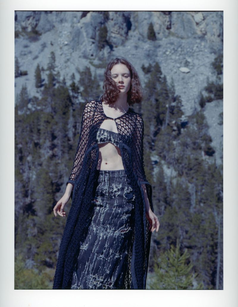 Woman poses in nature, wearing a matching skirt and top with a sheer black crocheted shawl, set against a mountain and forest backdrop.