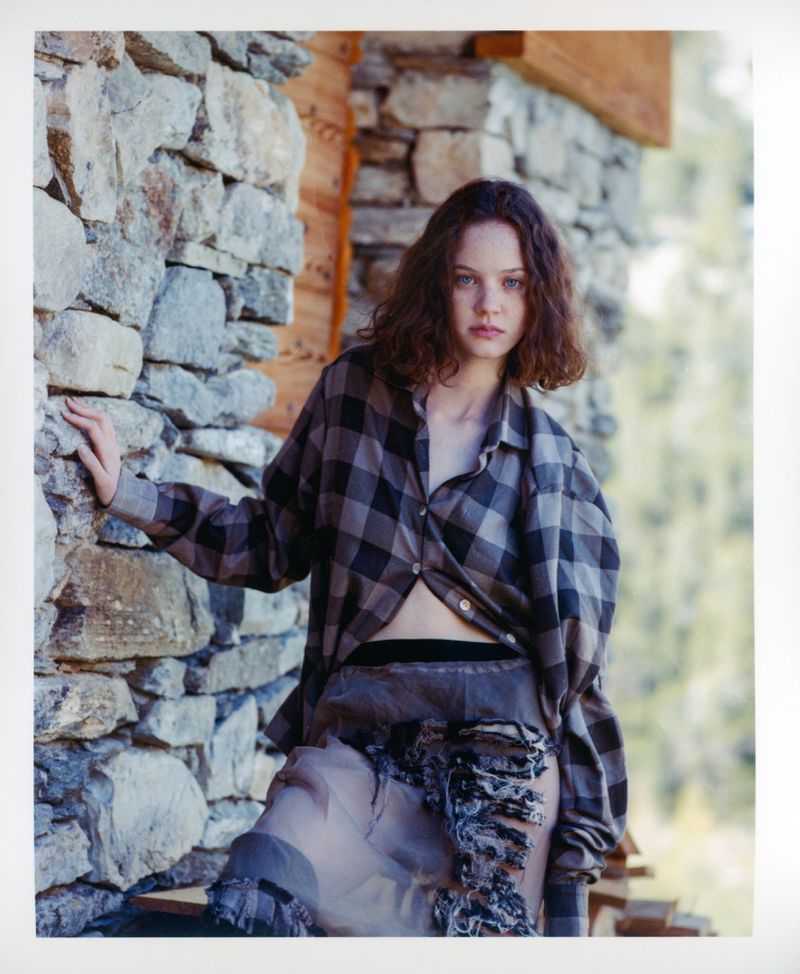 Young woman poses against a stone wall wearing a grey plaid shirt and flowing skirt in an outdoor setting with natural light.