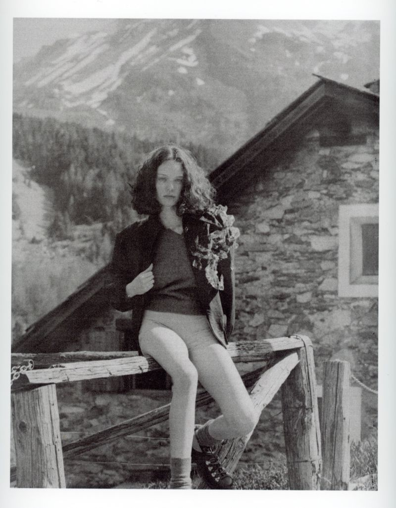 Monochrome shot of woman sitting on wooden fence with stone building backdrop and snow-capped mountain background in an outdoor setting.