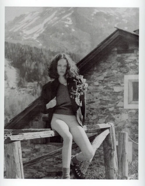 Monochrome shot of woman sitting on wooden fence with stone building backdrop and snow-capped mountain background in an outdoor setting.