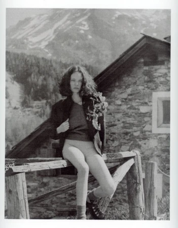 Monochrome shot of woman sitting on wooden fence with stone building backdrop and snow-capped mountain background in an outdoor setting.