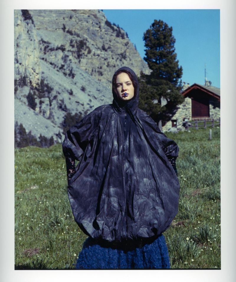 Fashion editorial shot of a young woman in an avant-garde poncho posing in a mountain meadow against a backdrop of a rustic cabin and mountain.