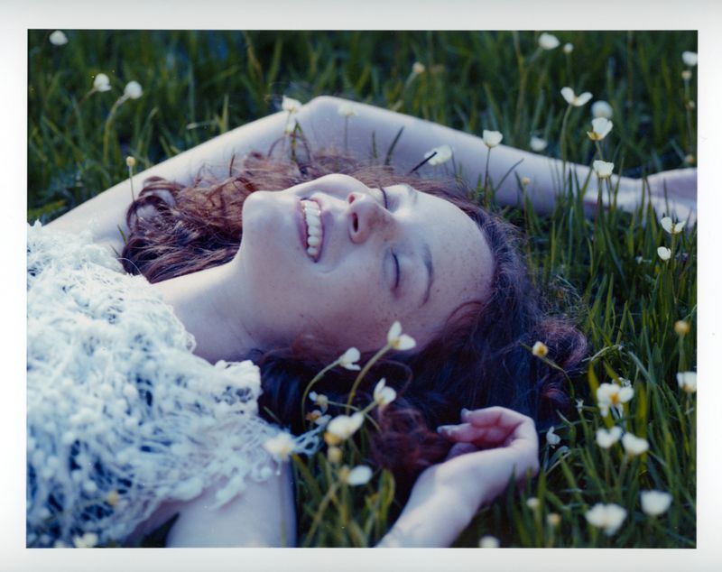 A smiling young woman with curly brown hair lying back on a grassy field surrounded by small white wildflowers.