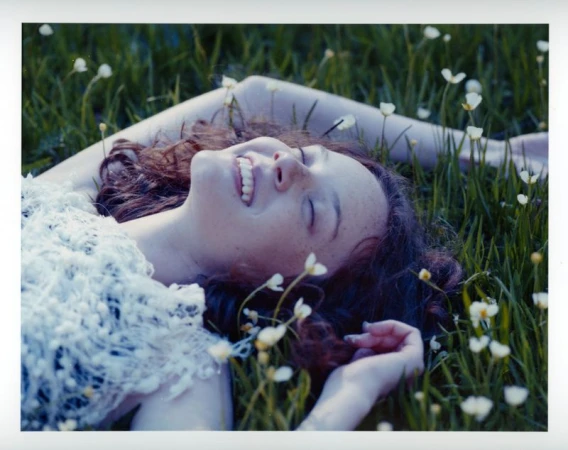 A smiling young woman with curly brown hair lying back on a grassy field surrounded by small white wildflowers.