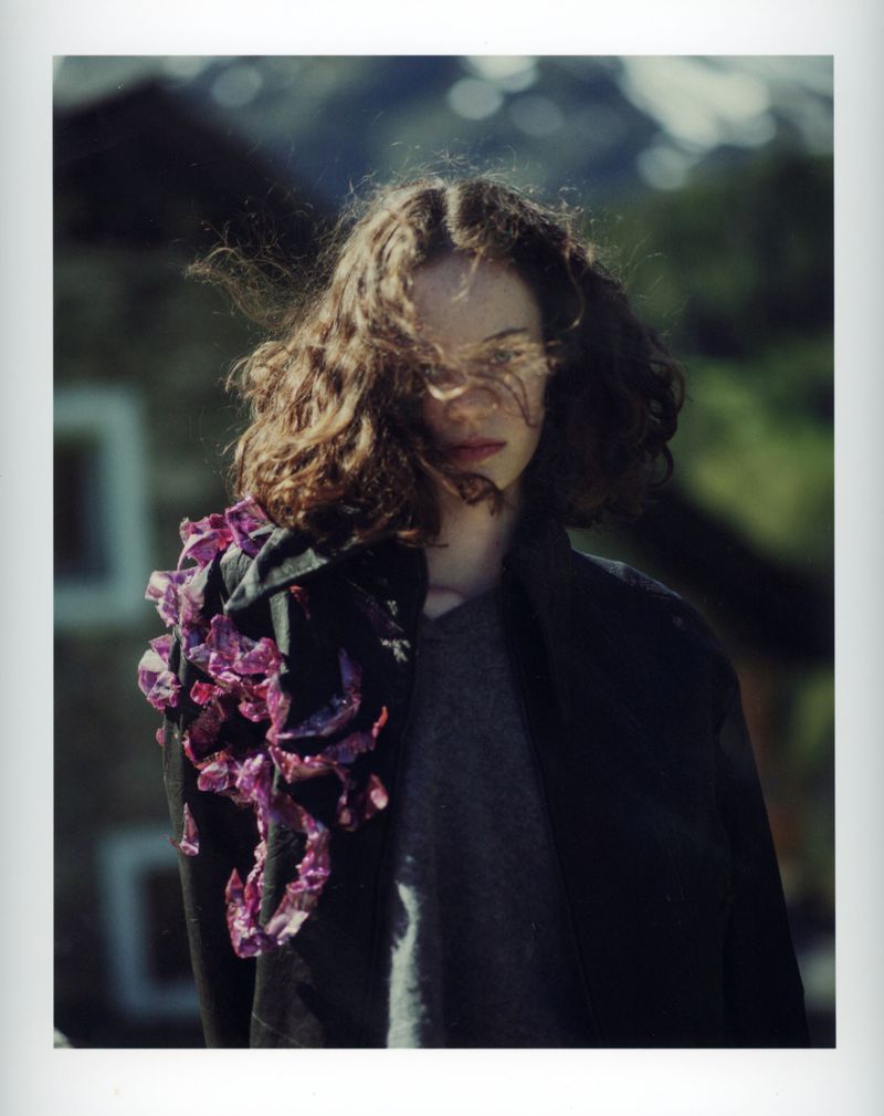 Young woman with curly hair stands outdoors wearing a black coat adorned with magenta textile flowers, set against blurred foliage.