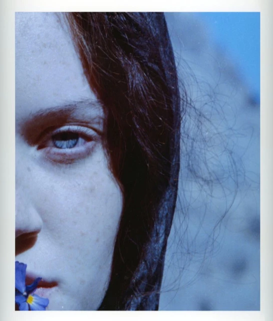 Close-up studio portrait of a dark-haired, light-skinned woman with a blue flower in her mouth, set against a blue gradient background.