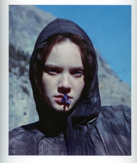 Close-up portrait of a young woman in a dark hooded garment, holding a small purple flower in her mouth, against a background of blue sky and mountains.