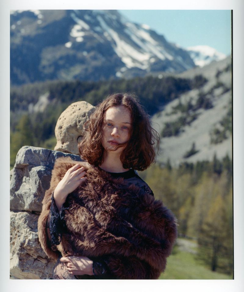 Woman in a fur jacket poses near a stone wall in a mountain landscape, bathed in natural light. She gazes at the camera, creating a serene scene.