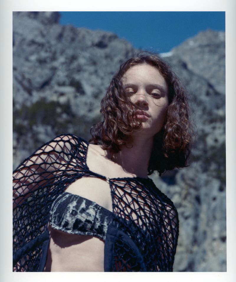 Portrait of a woman with auburn curly hair wearing a black net garment, posing in front of a background featuring a mountain range and blue sky.