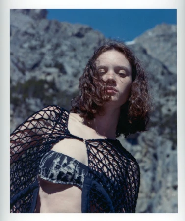 Portrait of a woman with auburn curly hair wearing a black net garment, posing in front of a background featuring a mountain range and blue sky.