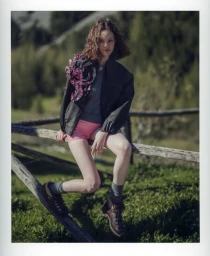 Woman wearing dark jacket with floral embellishment, pink shorts, and hiking boots sitting on a wooden fence outdoors.