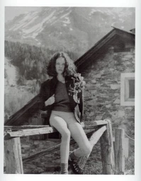 Black and white editorial portrait of a woman seated on a wooden fence before a stone alpine house and snowy mountain.