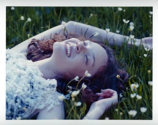 A smiling young woman with curly brown hair lying back on a grassy field surrounded by small white wildflowers.