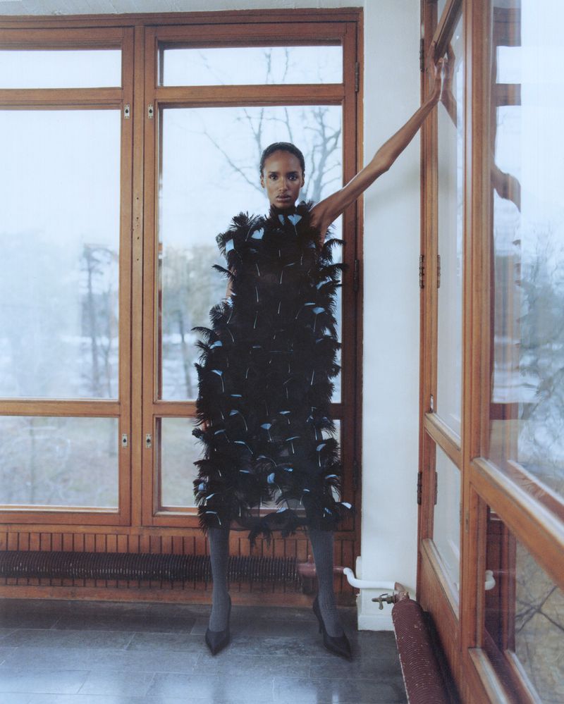 A slender woman strikes a pose indoors wearing a black feathered dress, grey tights, and black heels, against a wall of windows.