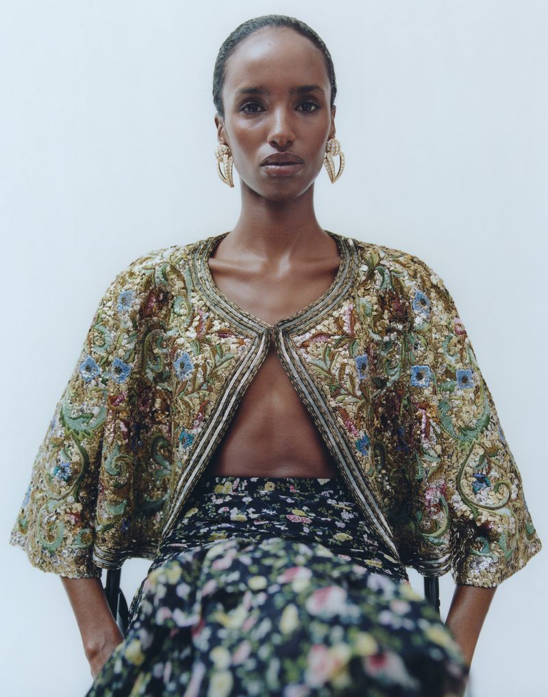Elegant woman with floral skirt and embroidered jacket posing in studio setting with soft light and wearing hoop earrings in close-up shot.
