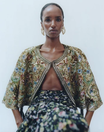 Elegant woman with floral skirt and embroidered jacket posing in studio setting with soft light and wearing hoop earrings in close-up shot.