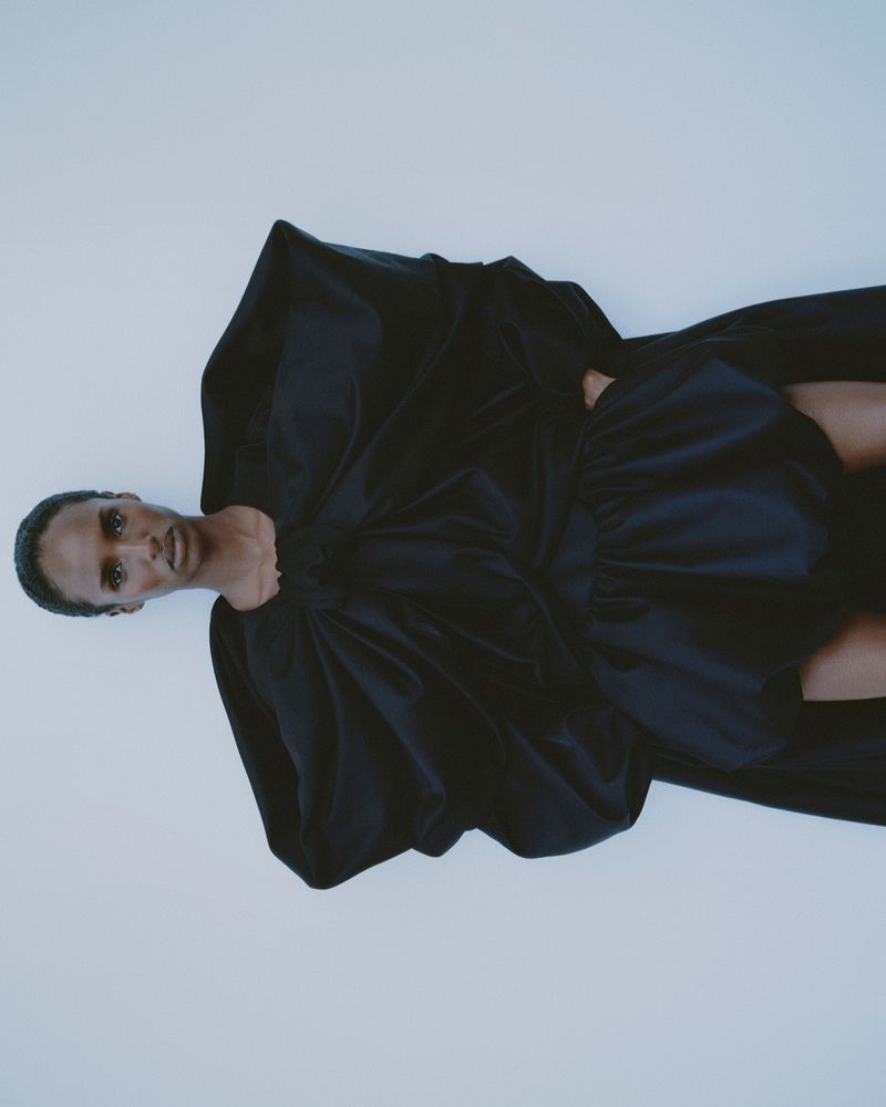 Fashion shot of a Black female model in a black gown with a dramatic bow posing against a white backdrop in a photo studio.