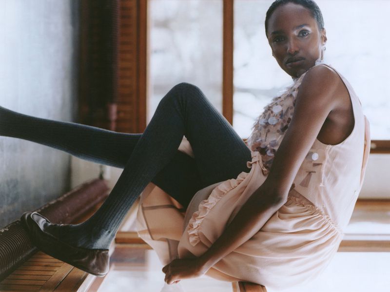 A woman poses in a sleeveless dress and leggings indoors, seated on a chair with her legs resting on a nearby structure, captured in a medium shot.