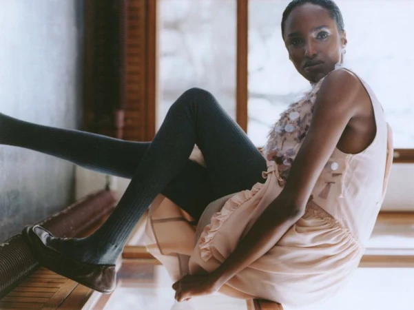 A woman poses in a sleeveless dress and leggings indoors, seated on a chair with her legs resting on a nearby structure, captured in a medium shot.