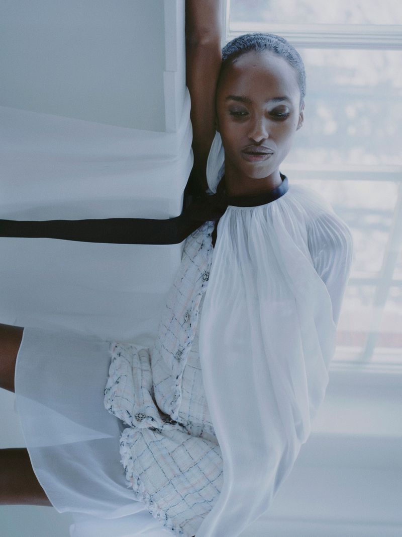 An African American female model poses indoors wearing a white and black dress near a window with soft lighting.