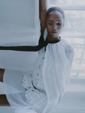 An African American female model poses indoors wearing a white and black dress near a window with soft lighting.