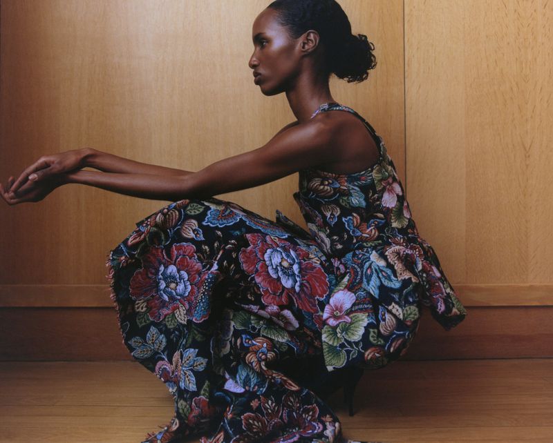 Adult woman in a patterned dress crouches in a studio setting. The portrait features a profile view with warm lighting and wood background.