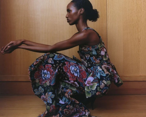 Adult woman in a patterned dress crouches in a studio setting. The portrait features a profile view with warm lighting and wood background.