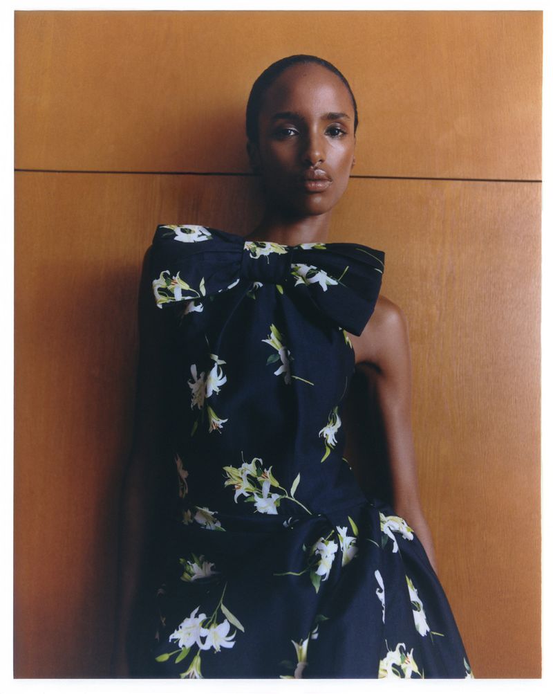 A Black woman with dark hair poses in a black floral dress with an oversized bow in a studio with tan wood paneling in the background.