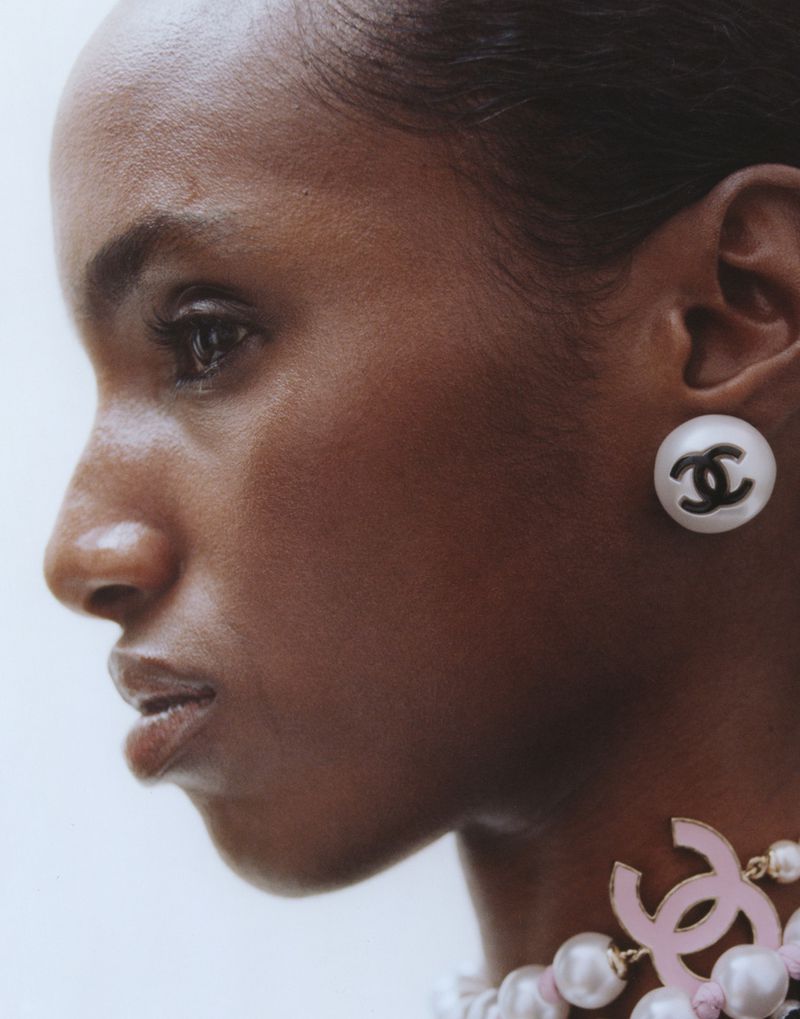 Profile shot of an adult woman with dark skin wearing pearl jewelry with a Chanel logo. She has dark eyes and is facing left.