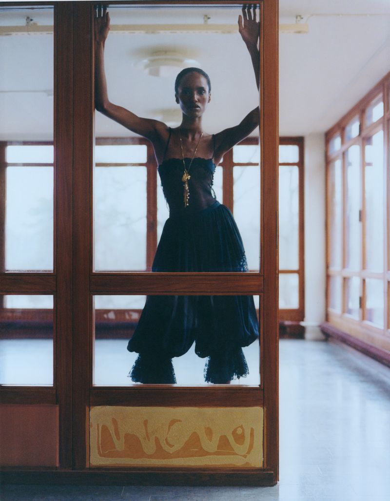 Fashionable woman in black dress poses between glass panes in minimalist setting with natural light and elegant long necklace.