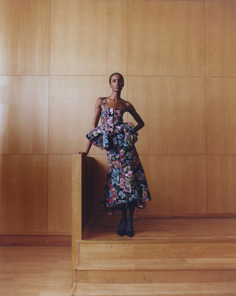 Elegant woman in floral couture dress posing against a wooden wall. Fashion shoot with high-end style and elegant indoor setting.