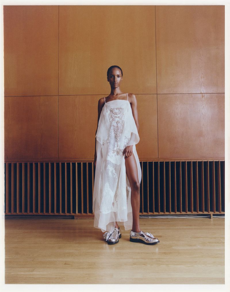 A full-length portrait of a slender woman in a white, semi-sheer dress standing in a minimalist interior with wood-paneled walls and floors.