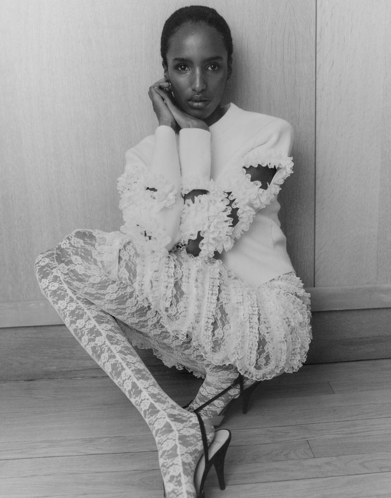 Black and white portrait of a woman in frilled top and lace leggings, seated and posing against a neutral wall. Fashion editorial.