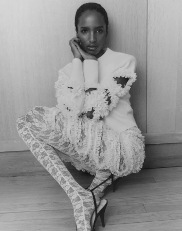 Black and white portrait of a woman in frilled top and lace leggings, seated and posing against a neutral wall. Fashion editorial.