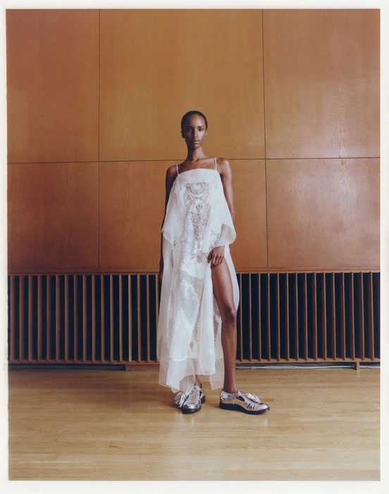 Model standing in a translucent white dress and silver shoes against a paneled wood wall for Violet Book fashion editorial