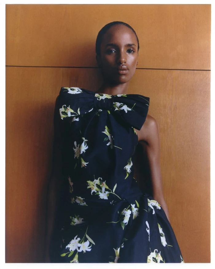 Model wearing a dark floral dress with a large bow detail against a warm wood panel wall.
