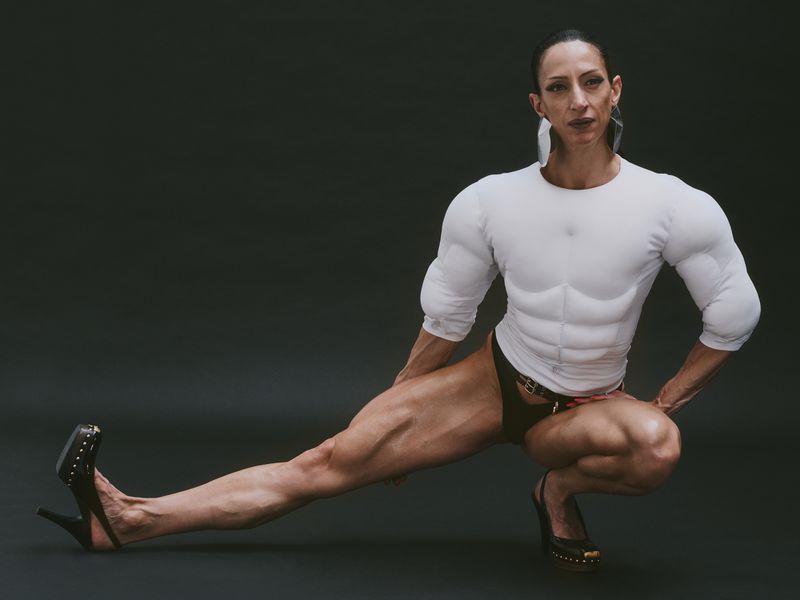Female bodybuilder in a white long-sleeve shirt and black stilettos in a studio. Focused fitness and fashion portrait.