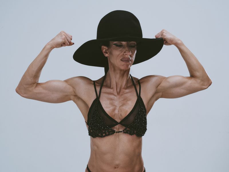 Muscular adult woman in black wide-brim hat and beaded bikini, flexing biceps against a neutral studio backdrop, showcasing strength and style.