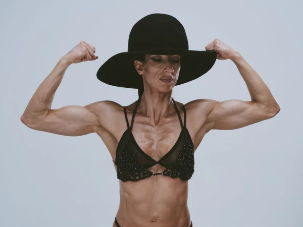 Muscular adult woman in black wide-brim hat and beaded bikini, flexing biceps against a neutral studio backdrop, showcasing strength and style.
