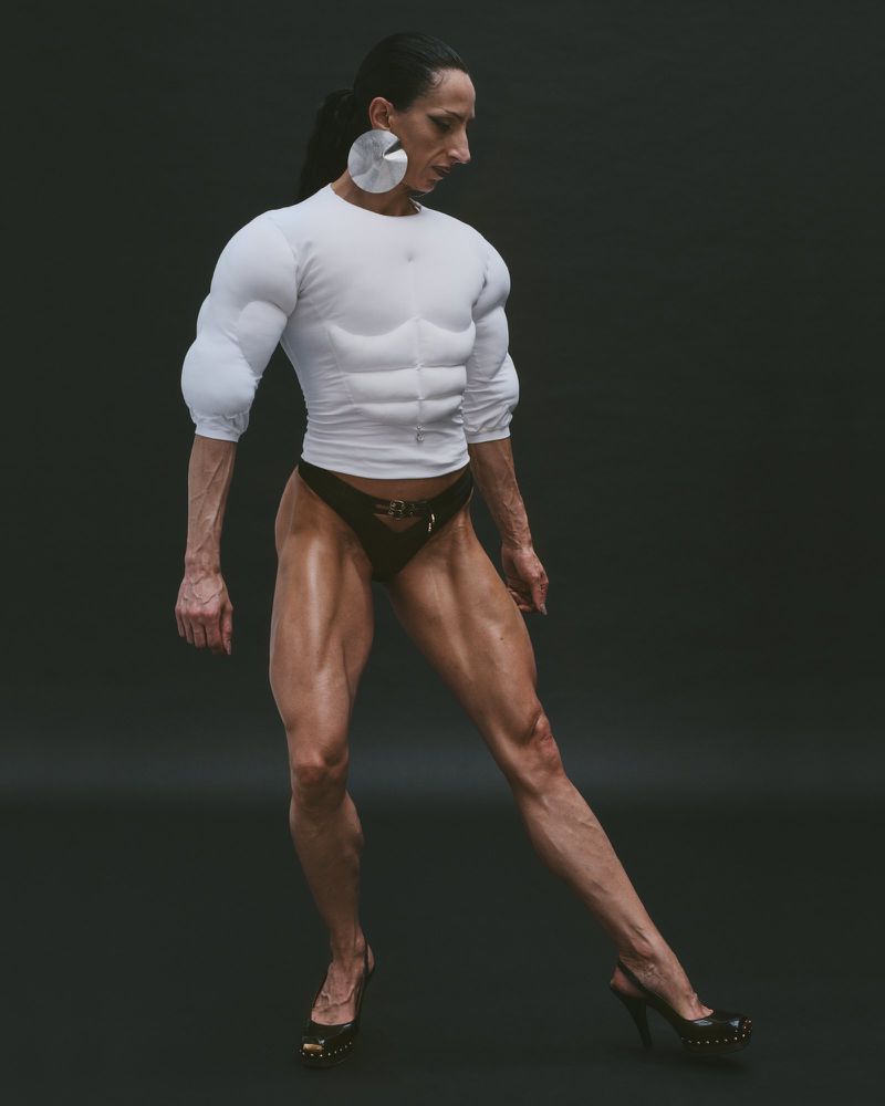 A muscular woman poses in a white top and black bikini bottoms against a dark grey background, highlighting fitness and body sculpting.