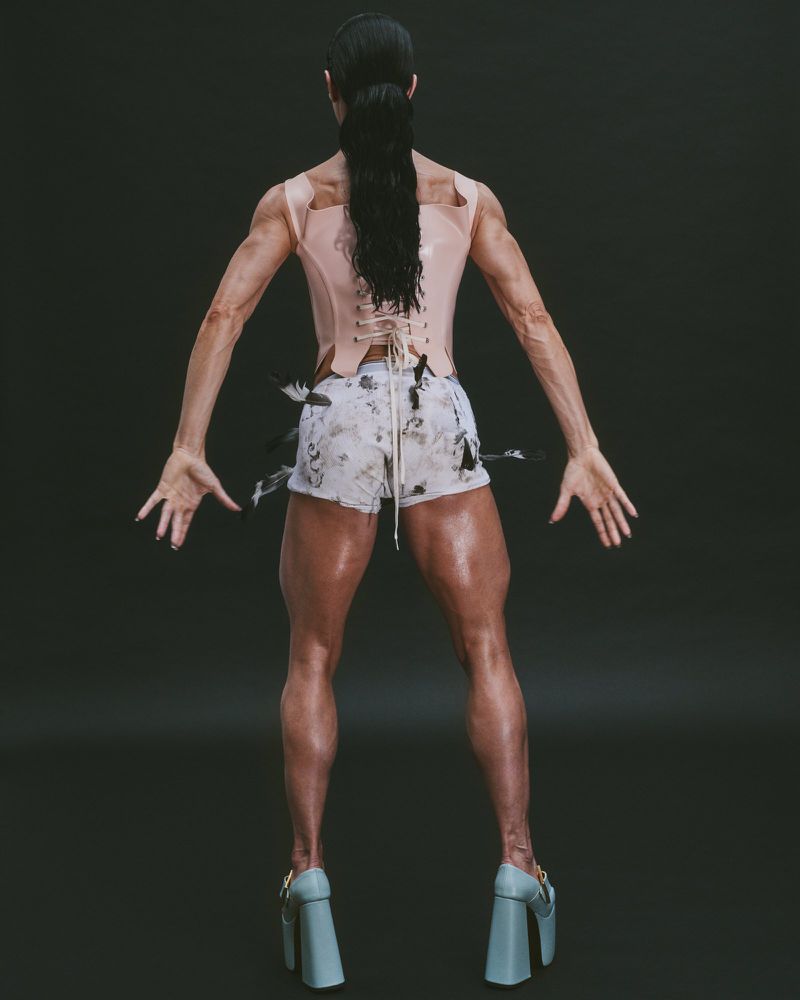 Back view of an adult female with dark hair, wearing a light pink corset top, patterned shorts, and high heels posing in a studio.